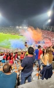Fuegos artificiales y banderas en estadio lleno durante partido de fútbol en Colombia.