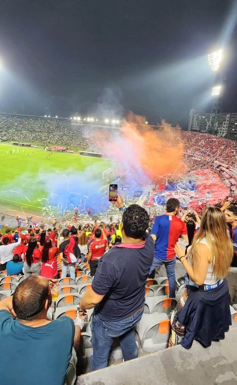 Fuegos artificiales y banderas en estadio lleno durante partido de fútbol en Colombia.